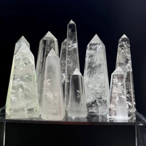 May include: A collection of clear quartz crystal obelisks of varying heights and shapes. The crystals are displayed on a reflective surface against a black background. The obelisks have pointed tops and visible internal structures.