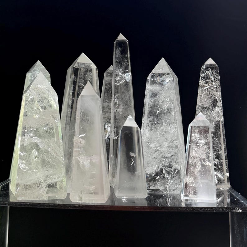 May include: A collection of clear quartz crystal obelisks of varying heights and shapes. The crystals are displayed on a reflective surface against a black background. The obelisks have pointed tops and visible internal structures.