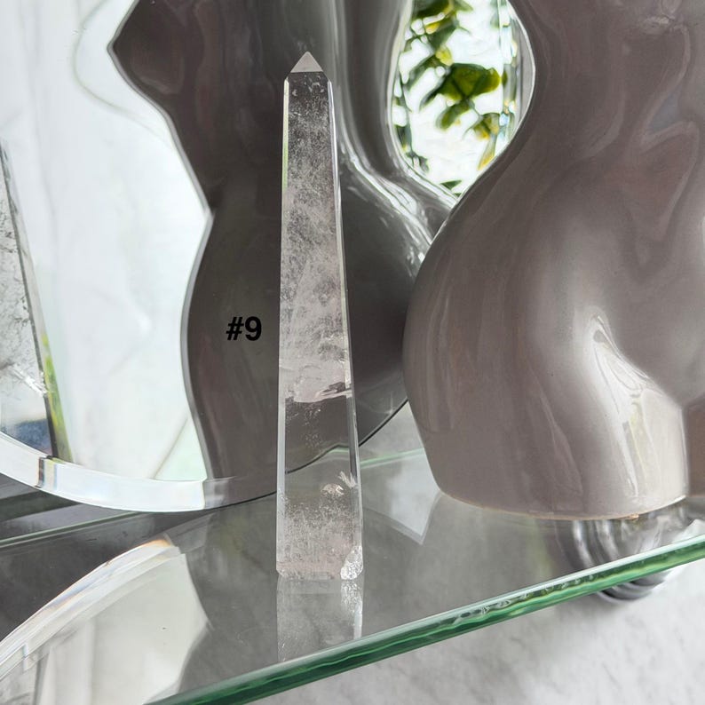 May include: A clear quartz crystal obelisk with a pointed top. The crystal is displayed on a glass shelf, with the number "#9" visible on the side. The background includes a blurred reflection and a decorative vase.