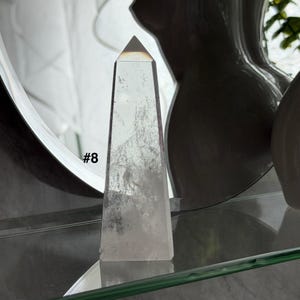May include: A clear quartz obelisk with a pyramidal top. The crystal has internal inclusions and is labeled with the number "#8". The obelisk is displayed on a glass shelf.