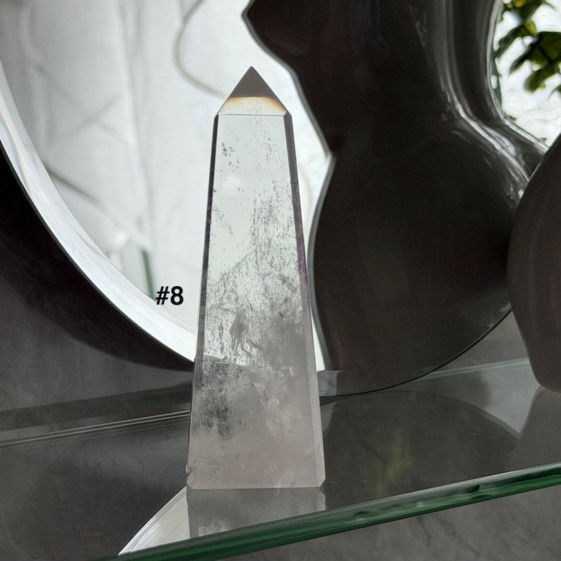 May include: A clear quartz obelisk with a pyramidal top. The crystal has internal inclusions and is labeled with the number "#8". The obelisk is displayed on a glass shelf.