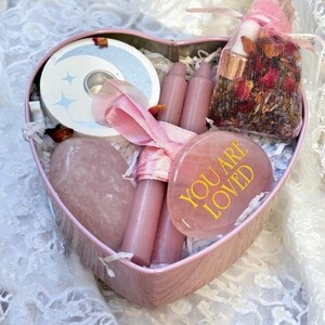May include: A pink heart-shaped box filled with self-care items. Inside are pink candles, a rose quartz heart, a stone with "YOU ARE LOVED" in gold lettering, a moon-themed candle, and a small bag of dried flowers and oils.
