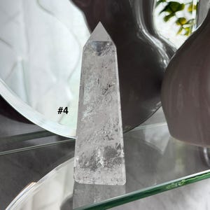 May include: A clear quartz crystal point with a faceted top. The crystal is rectangular with a tapered shape. The text "#4" is visible on the left side of the crystal. The crystal is displayed on a glass shelf.