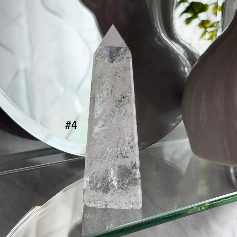May include: A clear quartz crystal point with a faceted top. The crystal is rectangular with a tapered shape. The text "#4" is visible on the left side of the crystal. The crystal is displayed on a glass shelf.