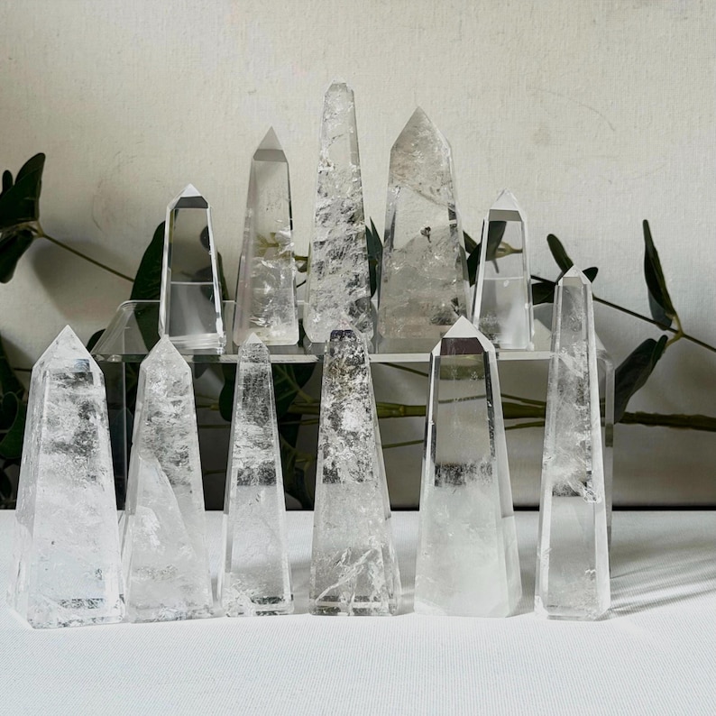 May include: A collection of clear quartz crystal obelisks of varying sizes. The crystals are arranged on a clear shelf and white surface, with green foliage in the background. The obelisks have pointed tops and are translucent.