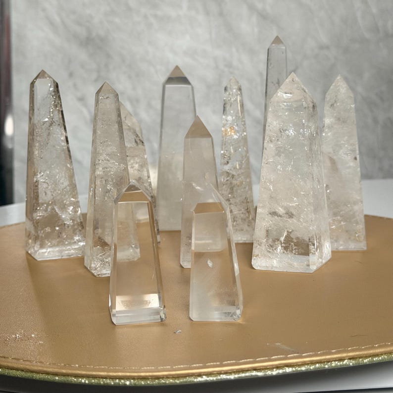 May include: A collection of clear quartz crystal obelisks of varying sizes and shapes. The crystals are arranged on a gold-colored tray, with a light-colored background. The obelisks have pointed tops and are translucent.