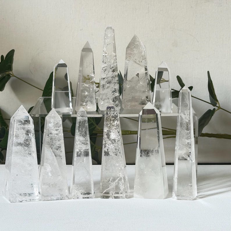 May include: A collection of clear quartz crystal obelisks of varying sizes and shapes. The crystals are arranged on a clear display stand and a white surface. The obelisks have pointed tops and are translucent, with visible internal patterns.