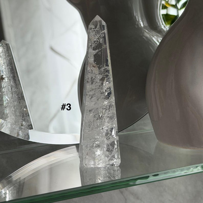 May include: A clear quartz crystal point with visible internal fractures. The crystal is shaped like a tall, slender obelisk. The text "#3" is visible on the crystal. The crystal is displayed on a glass shelf.