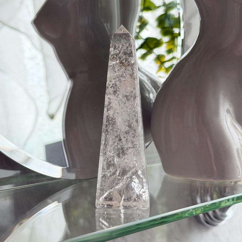 May include: A clear quartz crystal obelisk stands on a glass shelf. The crystal is a light, translucent color with internal textures. Two gray decorative vases are in the background. The obelisk is a decorative item.