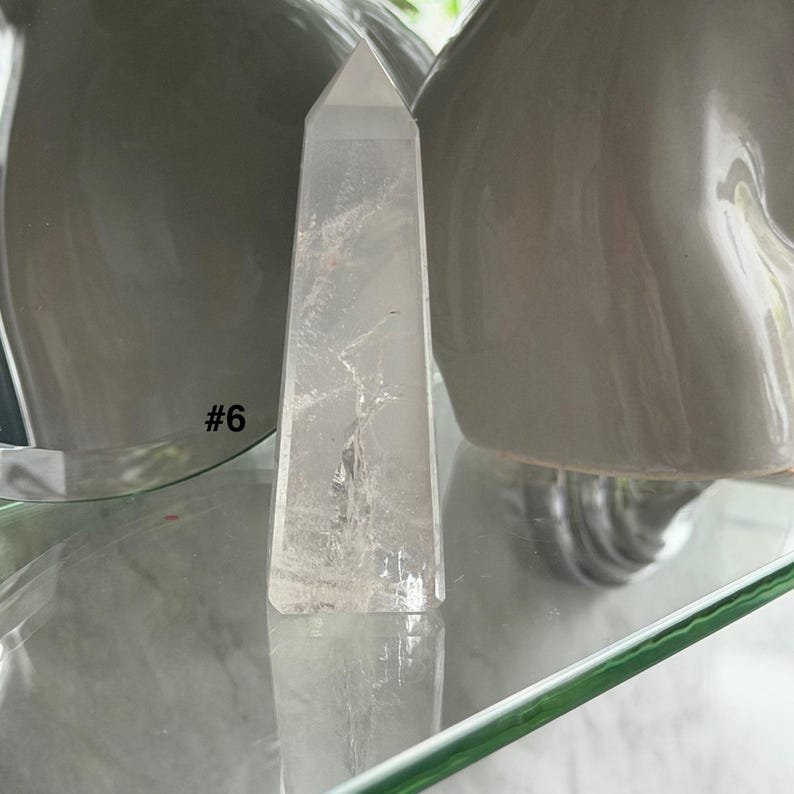 May include: A clear quartz crystal obelisk with a pointed top. The crystal is displayed on a glass shelf, with the number "#6" visible. The background includes two gray ceramic vases.