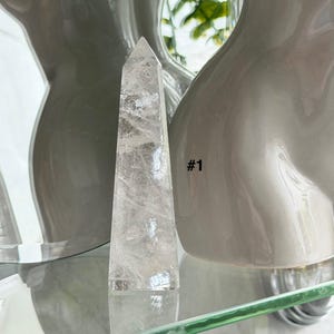 May include: A clear quartz crystal point with a pointed top. The crystal is standing upright on a glass shelf. The background includes a white ceramic vase and the text "#1".