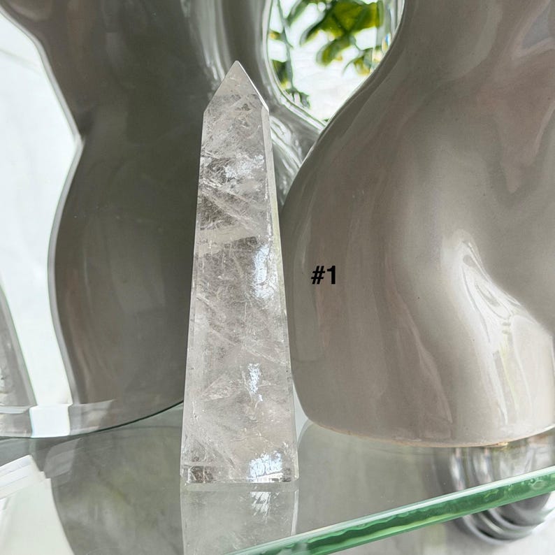 May include: A clear quartz crystal point with a pointed top. The crystal is standing upright on a glass shelf. The background includes a white ceramic vase and the text "#1".