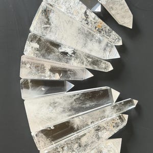 May include: Clear quartz crystal points arranged on a dark surface. The crystals are transparent with visible internal textures and sharp, pointed ends. The arrangement suggests a collection of natural, raw gemstones.