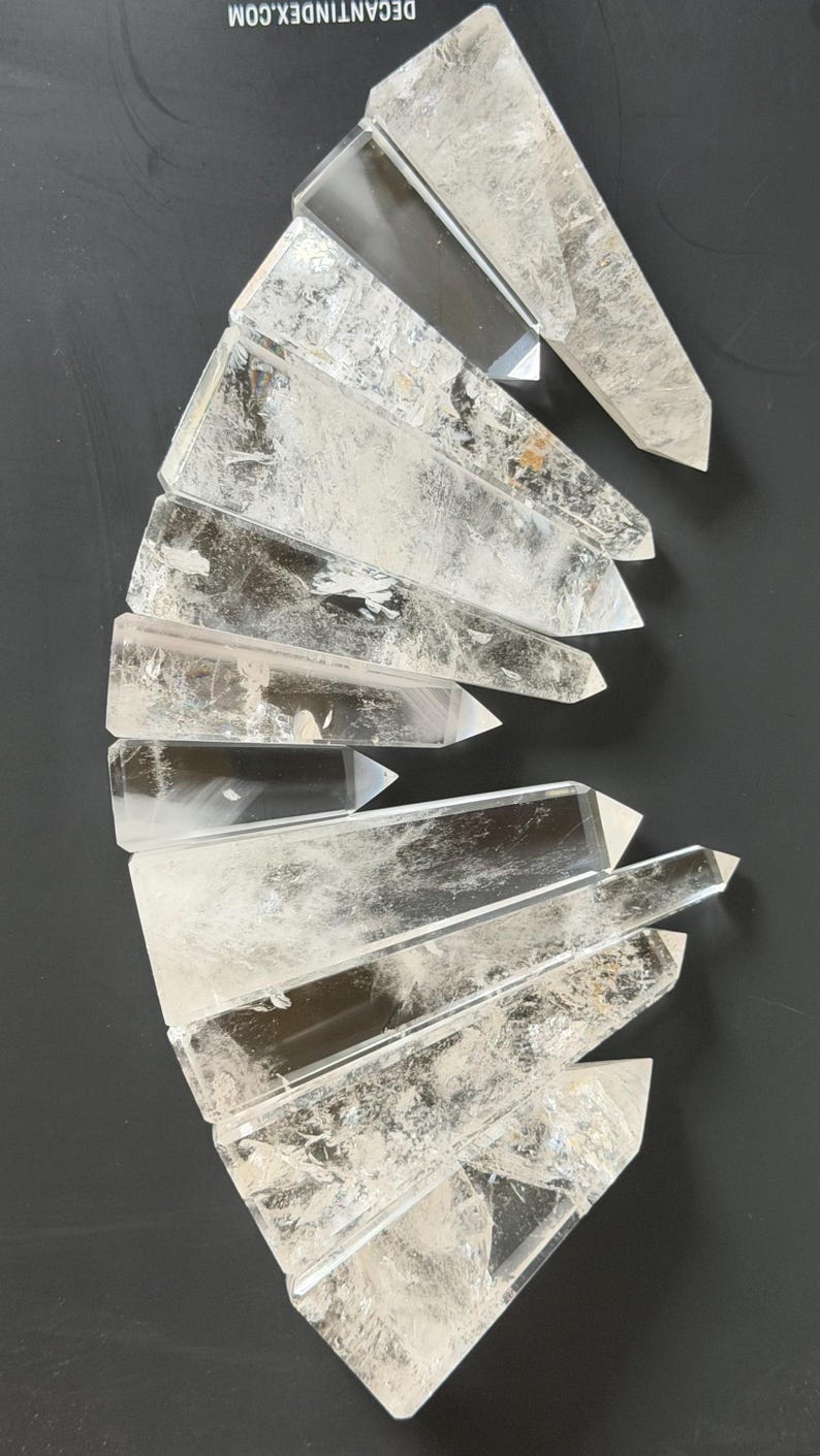 May include: Clear quartz crystal points arranged on a dark surface. The crystals are transparent with visible internal textures and sharp, pointed ends. The arrangement suggests a collection of natural, raw gemstones.