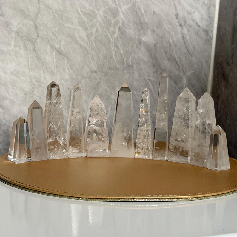 May include: A collection of clear quartz crystal points of varying heights and shapes. The crystals are arranged in a row on a gold-colored surface, with a light gray marble-like background. The crystals are transparent and reflect light.