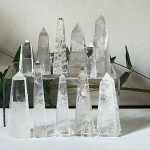May include: A collection of clear quartz crystal obelisks of varying sizes. The crystals are arranged on a clear acrylic display stand and a white surface, with green foliage in the background. The obelisks have pointed tops and visible internal structures.