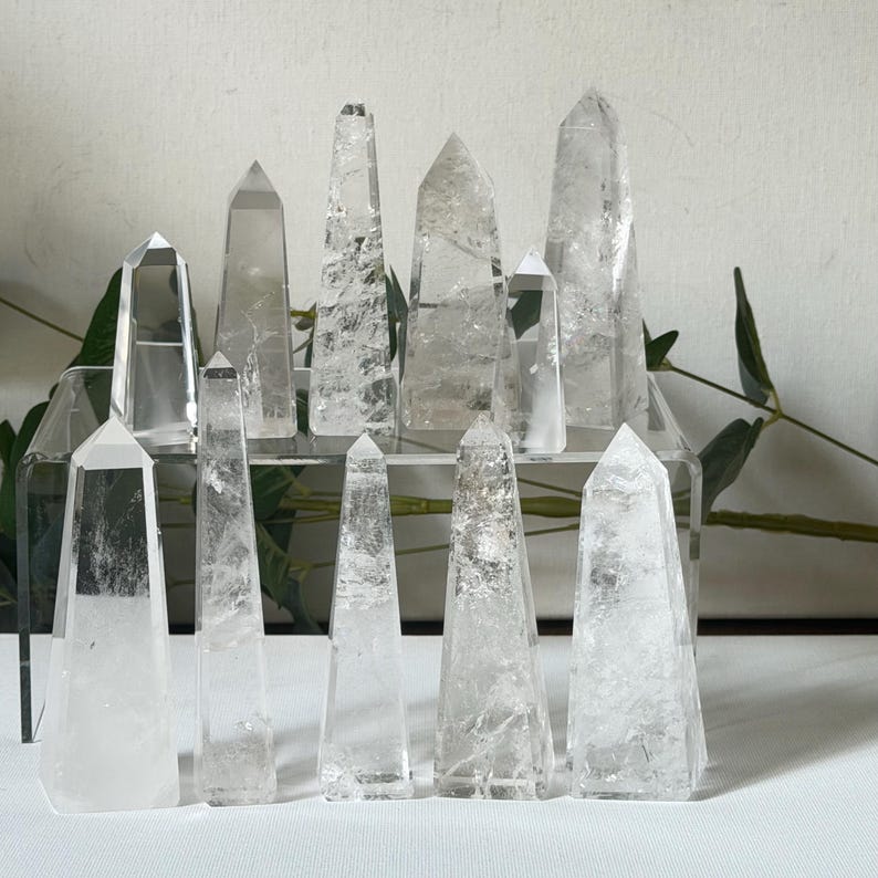 May include: A collection of clear quartz crystal obelisks of varying sizes. The crystals are arranged on a clear acrylic display stand and a white surface, with green foliage in the background. The obelisks have pointed tops and visible internal structures.