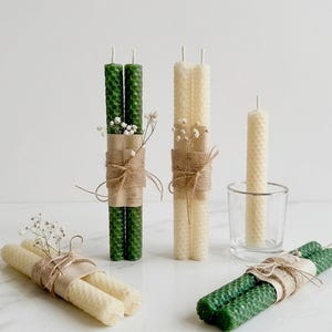 May include: An assortment of decorative candles in various shades of green and cream. The candles are bundled in pairs and decorated with twine and small sprigs of white flowers. One candle is in a clear glass.