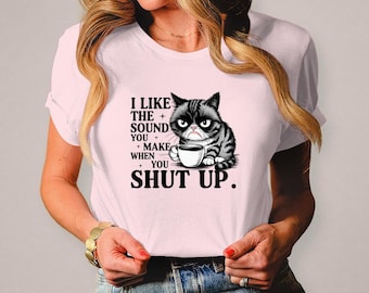 May include: A light pink t-shirt featuring a grumpy cat illustration holding a coffee cup. The text on the shirt reads "I LIKE THE SOUND YOU MAKE WHEN YOU SHUT UP."
