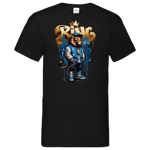Può includere: T-shirt nera con un leone dei cartoni animati che indossa una giacca, un berretto e scarpe da ginnastica blu. Il leone è in piedi davanti a graffiti dorati che recitano "King". Il design include simboli del dollaro e una corona.