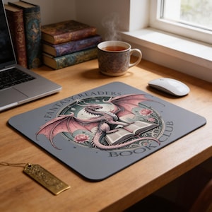 Könnte beinhalten: Ein rechteckiges Mauspad mit einer kreisförmigen Illustration eines rosa Drachen, der auf einem offenen Buch sitzt, umgeben von floralen Akzenten und dem Text FANTASY READERS BOOK CLUB, auf grauem Hintergrund mit abgerundeten Ecken.