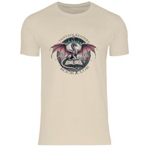 Könnte beinhalten: Ein beiges T-Shirt mit einem Aufdruck eines rosa Drachen, der auf einem geöffneten Buch sitzt. Der Aufdruck ist von einem Kreis mit dem Text "Fantasy Readers Book Club" umgeben.