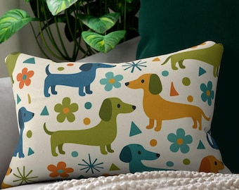 May include: A white throw pillow with a colorful pattern of dachshunds, flowers, and geometric shapes. The pillow is on a light gray couch with a white throw blanket.