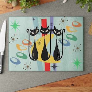May include: A blue and green retro-style kitchen cutting board with three black cats and a yellow crescent moon. The design features colorful abstract shapes and stars.