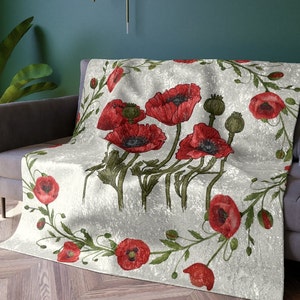 Peut inclure: Plaid blanc avec un motif de coquelicots rouges et de feuilles vertes. Le motif floral est encadré par une bordure de petites fleurs de pavot et de feuillage. Le plaid est drapé sur un canapé gris, un élément décoratif pour la maison.
