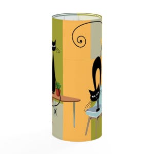 May include: A cylindrical lamp shade featuring a graphic illustration of two black cats on a background of orange, olive green, and light blue panels. One cat sits on a brown table, while the other arches its back on a light blue chair.