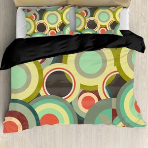 May include: Bedding set featuring a colorful, retro-style design. The duvet cover and pillowcases display overlapping circles in shades of green, yellow, brown, and red. A black comforter covers the top of the bed, creating a contrast with the patterned bedding.