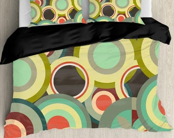 May include: Bedding set featuring a colorful, retro-style design. The duvet cover and pillowcases display overlapping circles in shades of green, yellow, brown, and red. A black comforter covers the top of the bed, creating a contrast with the patterned bedding.