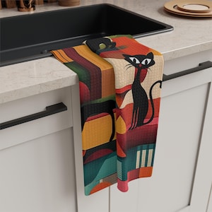 May include: A decorative kitchen towel featuring a black cat with large eyes and a long tail. The towel has a colorful geometric design with red, orange, green, and yellow hues. The towel is draped over a kitchen sink.
