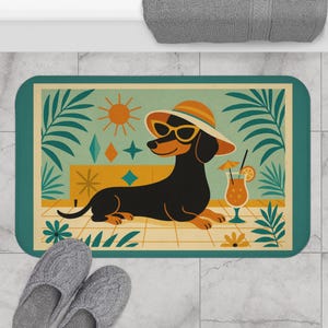 May include: A rectangular bath mat featuring a cartoon dachshund wearing sunglasses and a sun hat, lounging by a cocktail. The mat has a teal border and a retro beach scene design with a sun, palm fronds, and geometric shapes.