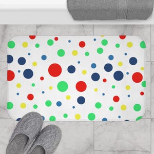 May include: A rectangular white bath mat with a colorful polka dot pattern. The dots are in various sizes and colors, including red, green, yellow, and blue. The mat is placed on a tiled floor, with a pair of gray slippers visible.
