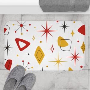 May include: A rectangular bath mat featuring a white background with a repeating pattern of red and yellow geometric shapes, including triangles, diamonds, and starbursts, accented by thin black lines and small black and yellow dots.