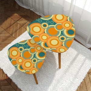 May include: Two retro-style side tables with a triangular top and wooden legs. The table tops feature a colorful pattern of overlapping circles in shades of orange, yellow, and teal. The tables are placed on a white rug.