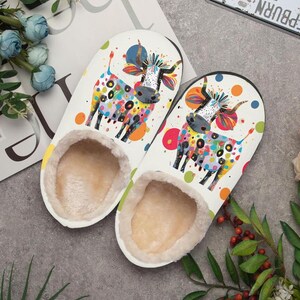 May include: A pair of white slippers with a colorful cow design. The cow is decorated with polka dots and rainbow colors. The slippers have a soft, beige interior.