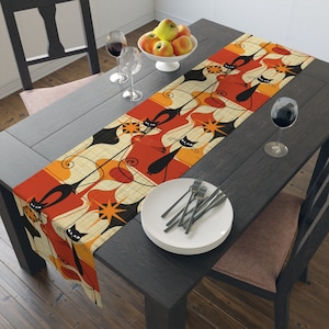 May include: A black and orange table runner with a retro design featuring stylized black cats and orange and yellow geometric shapes. The runner is on a dark wood table with a place setting.