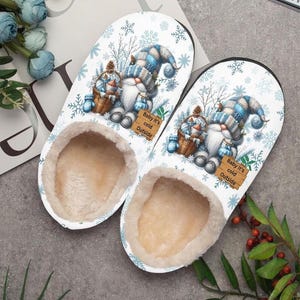 Puede incluir: Un par de zapatillas blancas con un dise&ntilde;o de gnomo de invierno. Las zapatillas presentan un gnomo con un gorro a cuadros azul y blanco, sosteniendo una cesta. El texto "Baby it's cold Outside" est&aacute; en un letrero de madera. El interior est&aacute; forrado con un material beige suave.