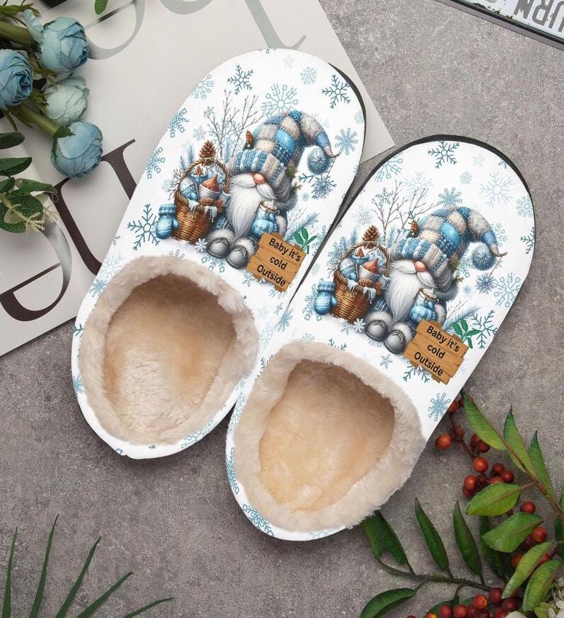 Puede incluir: Un par de zapatillas blancas con un dise&ntilde;o de gnomo de invierno. Las zapatillas presentan un gnomo con un gorro a cuadros azul y blanco, sosteniendo una cesta. El texto "Baby it's cold Outside" est&aacute; en un letrero de madera. El interior est&aacute; forrado con un material beige suave.