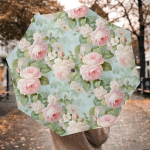 May include: A floral umbrella featuring a pattern of pink and white roses with green leaves against a light blue background. The umbrella is open, showcasing the detailed design and a black button at the center.