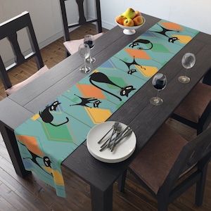 May include: A blue and yellow table runner with a geometric pattern and black silhouettes of cats. The runner is on a dark wood table with two chairs and wine glasses.