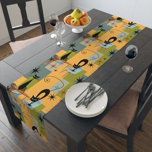 May include: A retro-style table runner featuring a repeating pattern of black cat illustrations on a mid-century modern design. The runner has a color palette of orange, green, and blue, with starburst and geometric shapes. The runner is placed on a dark wooden table.