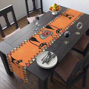 May include: Orange table runner with a geometric pattern featuring black cats, stars, and a retro design. The runner is on a dark wood table with two chairs and a glass of wine.