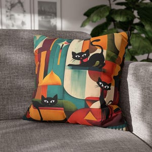 Puede incluir: Cojín cuadrado con un diseño abstracto colorido con ilustraciones de gatos negros. El cojín tiene una mezcla de tonos turquesa, naranja, rojo y amarillo, con un patrón geométrico. Los gatos están en varias poses.