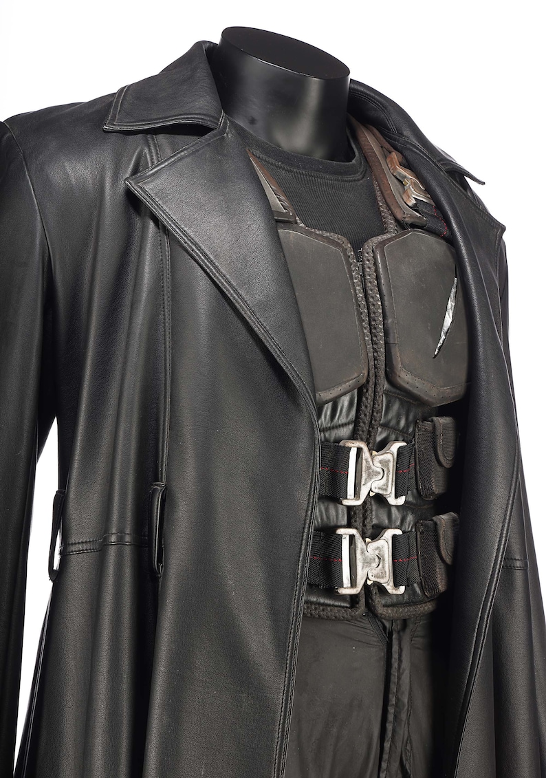 May include: A black leather trench coat with a black vest underneath. The vest has a silver buckle and straps.