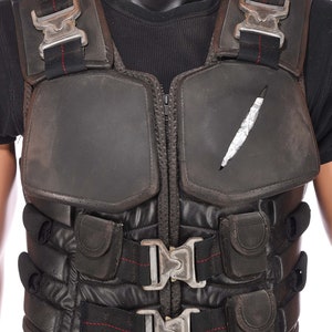 May include: Black tactical vest with multiple straps and buckles. The vest has a zipper closure and a padded front panel. There is a small tear in the front panel.