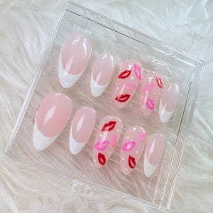 Puede incluir: Un conjunto de diez u&ntilde;as postizas de color rosa y blanco con una punta blanca y un dise&ntilde;o de labios rojos y rosas en algunas de las u&ntilde;as.