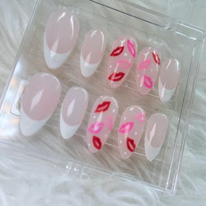 Puede incluir: Conjunto de diez u&ntilde;as postizas ovaladas con punta francesa blanca. Algunas u&ntilde;as tienen un dise&ntilde;o de labios rojos y rosas sobre fondo blanco.